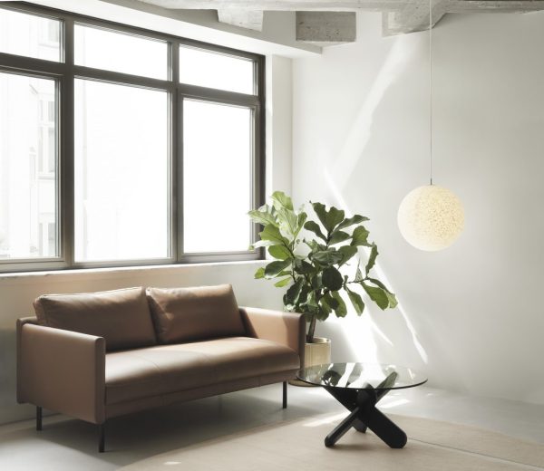 En moderne stue med en brun lædersofa, glas sofabord, stor potteplante og en NORMANN COPENHAGEN - Pix Pendel Ø30 hængelampe. Store vinduespartier giver naturligt lys, og vægge og loft er hvide med synlige bjælker.