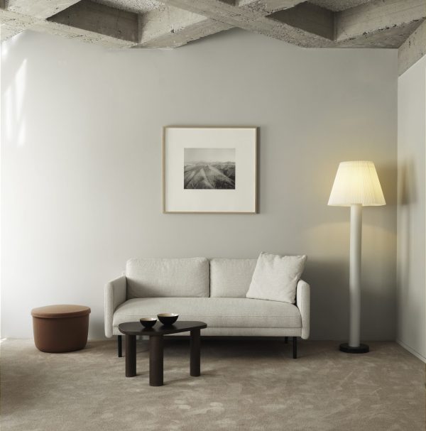 En moderne minimalistisk stue med en lysegrå sofa med en hvid hynde, et rundt brunt sofabord, en høj gulvlampe og en lille sidemøbler. Et indrammet landskabsfoto hænger over sofaen.