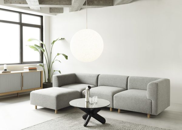 En minimalistisk stue med en NORMANN COPENHAGEN - Redo Modulsofa 110 Center, et rundt sort sofabord og et overdimensioneret sfærisk hængelys. Store vinduer, en potteplante og neutrale toner skaber et lyst, moderne rum.