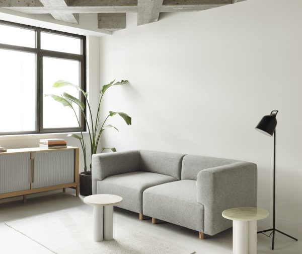 En minimalistisk stue med en grå NORMANN COPENHAGEN - Dit Bord, to runde sideborde, en gulvlampe og en stor potteplante ved siden af et vindue.