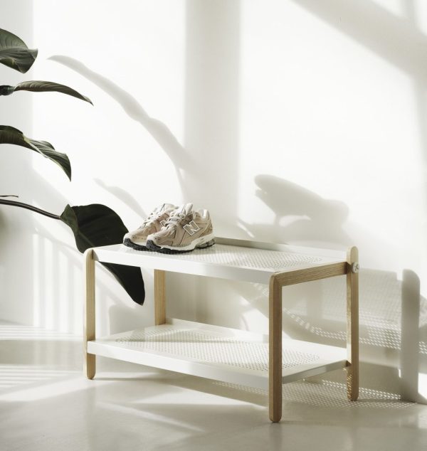 Et minimalistisk hvidt rum med en NORMANN COPENHAGEN - Sko Skostativ, der holder et par sneakers. sollys kaster bløde skygger på gulv og væg. en potteplante er til venstre, delvist i skygge.