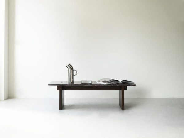 Et minimalistisk design med et mørkt træbord mod en almindelig hvid væg. Bordet rummer en metalkande, et klart glas og en stak bøger.