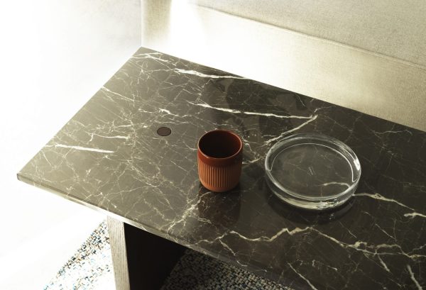 En keramisk kop og et glasaskebæger sidder på et poleret marmorbord nær en designmøbler sofa, fremhævet af naturligt sollys. Overfladen viser rig, mørk åring.