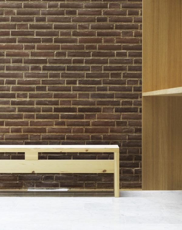 EN NORMANN COPENHAGEN Stretch Bænk 160 står op ad en murstensvæg i minimalistisk interiør, med hvidt gulv og et panel af lyst træ på højre side.