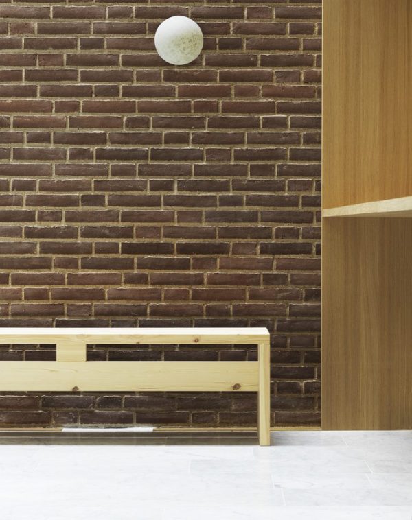 Et minimalistisk interiør med en rød murstensvæg med en NORMANN COPENHAGEN Pix Lampe Ø20, en enkel træbænk og en delvis udsigt til en trædør til højre. gulvet er lyst og glat.