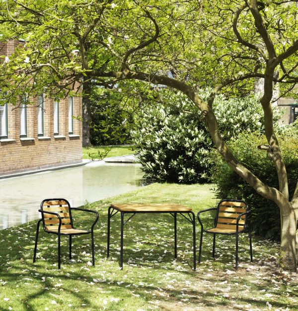 En rolig udendørs indstilling med en NORMANN COPENHAGEN - Vig Bord 90X80 og to stole under et blomstrende træ ved siden af en lille dam, med frodigt grønt græs og bygningens ydre i baggrunden.