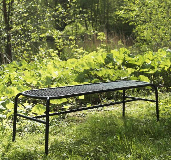 EN NORMANN KØBENHAVN - Vig Bænk sidder på grønt græs omgivet af frodigt løv i en solbeskinnet have. bænkens design har vandrette lameller og buede kanter.