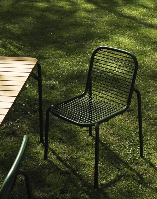 Et ensomt NORMANN COPENHAGEN - Vig Havestol med vandrette lameller står ved siden af et træbord, delvist støbt i skygge på et græsklædt område plettet med sollys.