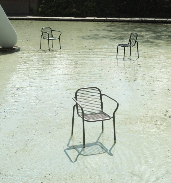 Tre NORMANN COPENHAGEN - Vig Havestols placeret på lavt vand, reflekterende på vandoverfladen, med sollys, der skaber mønstre på jorden. træer og en bygning er svagt synlige i baggrunden.