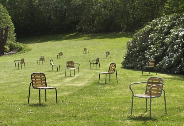 En solrig parkscene med flere tomme NORMANN COPENHAGEN - Vig Havestol-stole spredt tilfældigt hen over en velplejet græsplæne, omkranset af frodige grønne buske og træer.