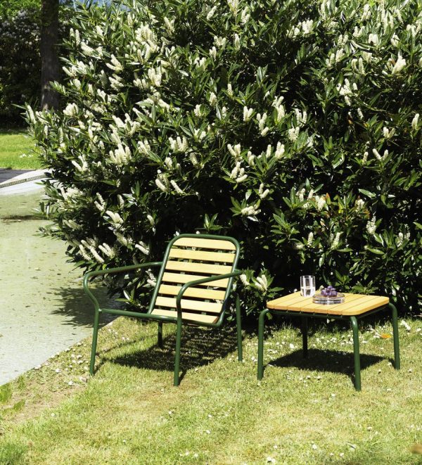 En grøn NORMANN COPENHAGEN - Vig Loungestol og et matchende lille bord ved siden af, sat på et græsareal med en frodig busk af hvide blomster i baggrunden under skarpt sollys.