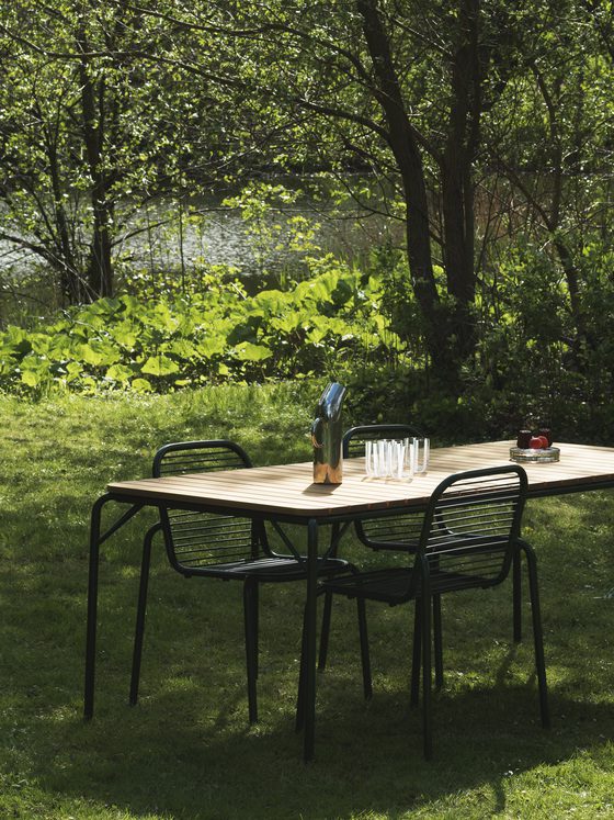 ET NORMANN COPENHAGEN - Vig Havestol spisebord sat med seks sorte stole i en solbeskinnet have, prydet med en blå kande, glas og en skål med jordbær, omgivet af frodigt grønt.