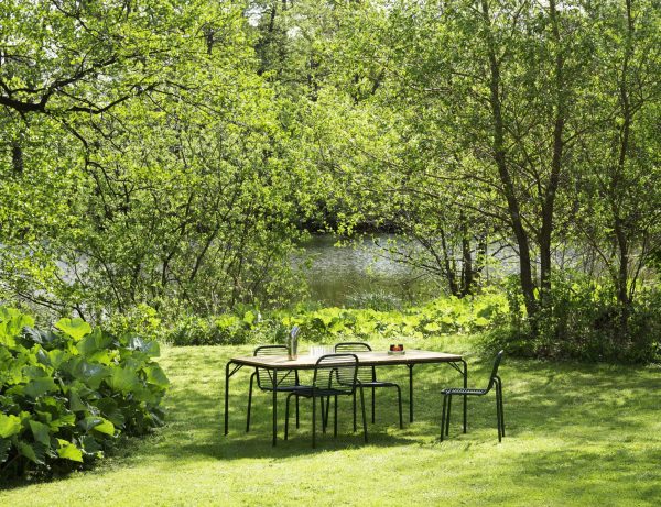 En fredfyldt have med et NORMANN COPENHAGEN - Vig Havestol spisebord og stole på græs med udsigt over en rolig dam, omgivet af frodige grønne træer og buske under en lys himmel.