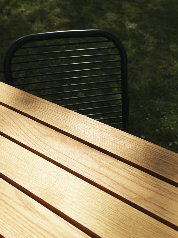 Nærbillede af en NORMANN COPENHAGEN - Vig Bord 90X200 med detaljeret træstruktur, der kaster skygger i sollys, med en del af en sort metalstol synlig i baggrunden, sat på en græsklædt overflade.