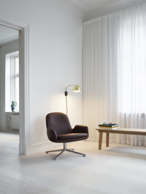 Et minimalistisk værelse med hvide vægge og trægulve med en elegant brun læderstol og en lille metalgulvlampe. Et træbord med bøger og et par grønne sneakers står tæt ved et vindue med gennemsigtige gardiner, og fremviser enkle, men funktionelle designmøbler.