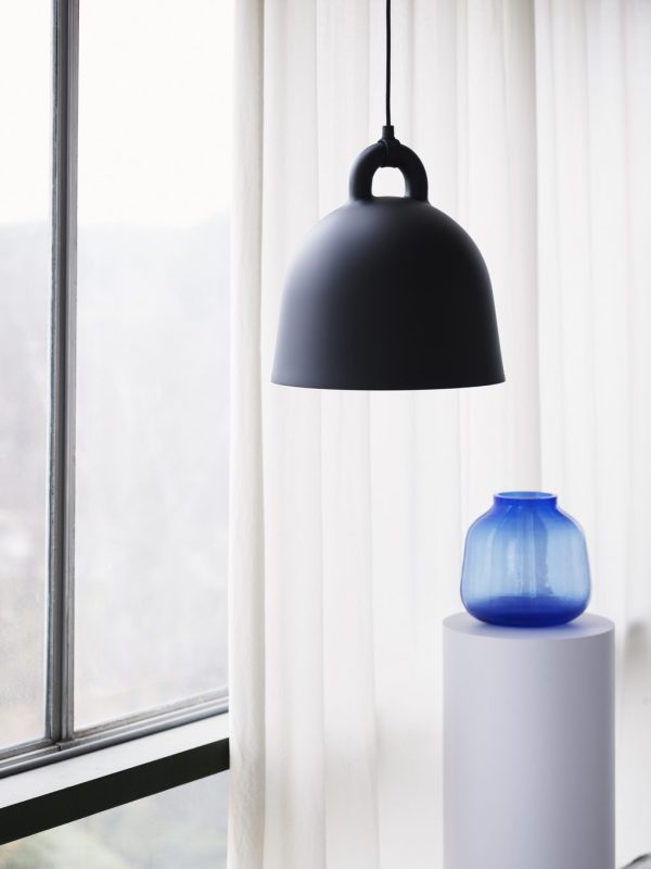 En matsort NORMANN COPENHAGEN - Bell Lampe Ø42 hænger nær et vindue med gennemsigtige gardiner, med udsigt over et sløret naturlandskab. ved siden af, på en hvid piedestal, står en lys blå vase.