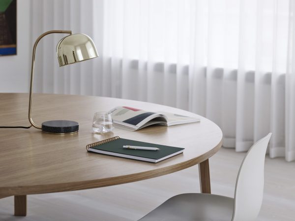 Et pænt organiseret studiebord med tændt messingbordslampe, magasin, notesbog, kuglepen og et klart glas vand, ledsaget af en designmøbler stol, i et rum med rene gardiner.