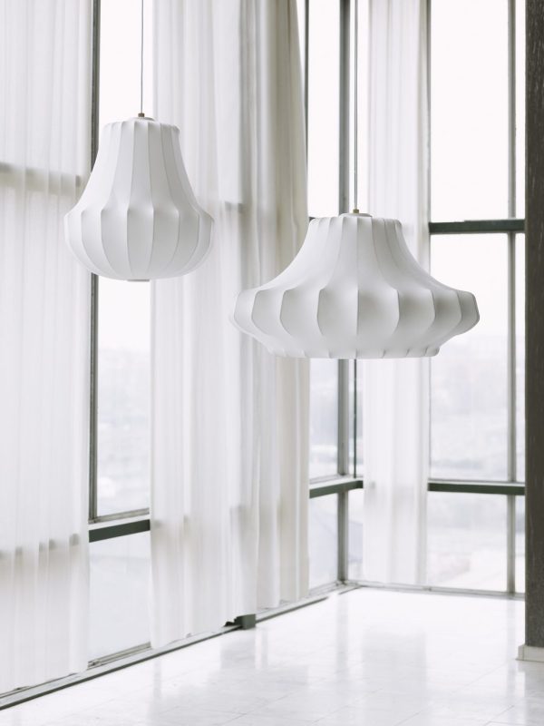 To moderne hvide pendellamper fra TomorrowsDesign, der hænger foran gulv-til-loft vinduer med gennemsigtige gardiner, med udsigt over et lyst, sløret bybillede.