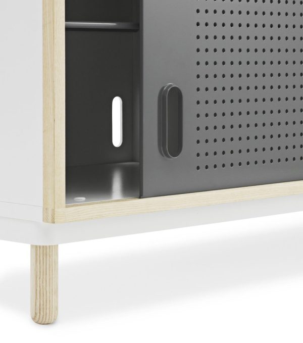 Nærbillede af en NORMANN COPENHAGEN - Kabino Skænk med et slankt design, med perforerede ventilationsåbninger, en lodret slids og et håndtag, alt sammen monteret på en hvid hylde med træben.