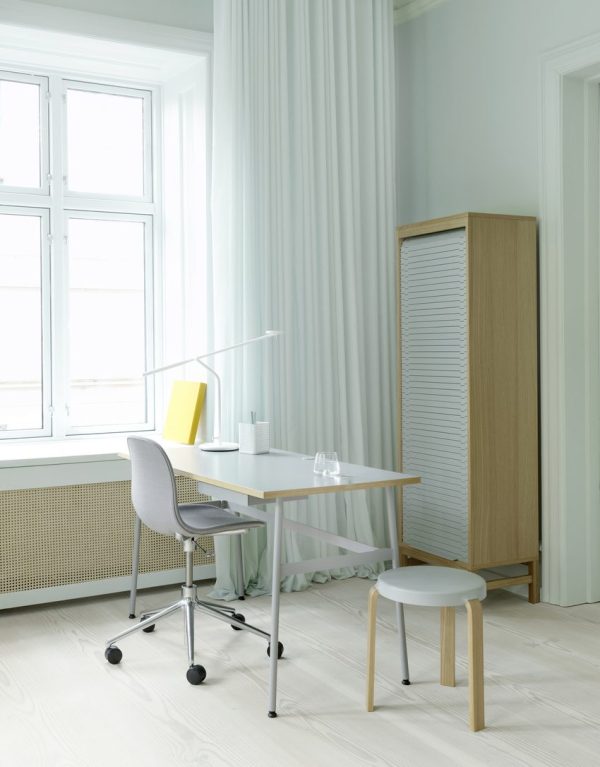 Et lyst, minimalistisk hjemmekontor med designmøbler med hvidt skrivebord og stol, en gul bog, hvid skrivebordslampe og en skammel. Et stort vindue og højskab er også synlige i et rum med lysegrønne vægge og trægulve.