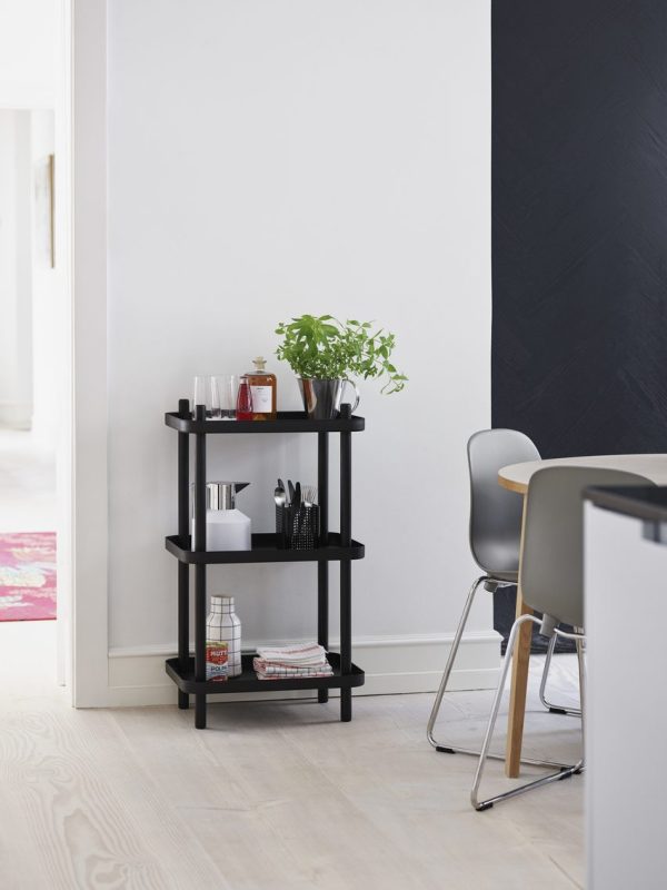 EN NORMANN COPENHAGEN - Block Reol står nær en hvid døråbning, fyldt med planter, bøger og køkkenting. en del af en spiseplads med en grå stol og et hvidt bord ses til højre.