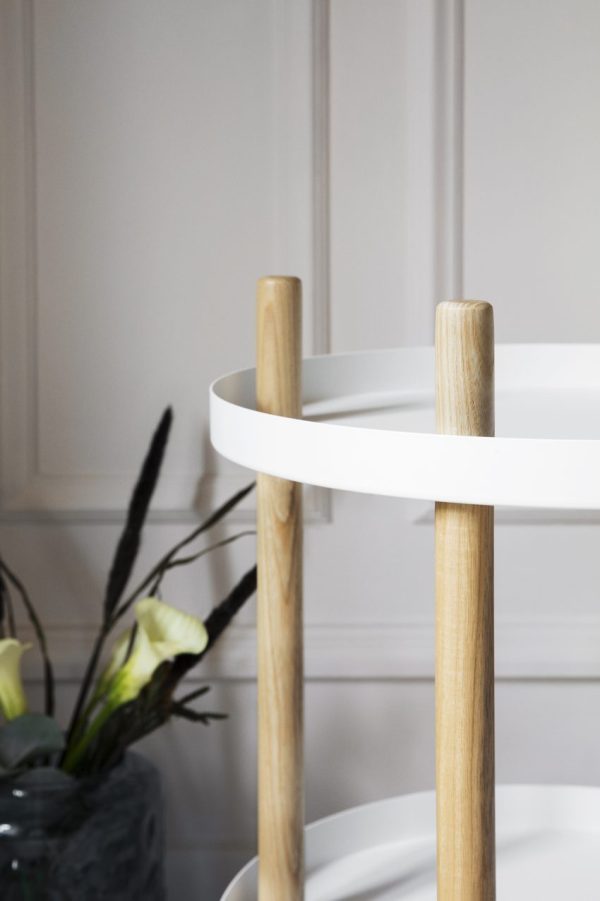 Nærbillede af en moderne hvid cirkulær vogn med naturlige træhåndtag, med TomorrowsDesign-æstetik, sat op mod en hvid panelvæg. En del af en mørk vase med blomster er synlig i baggrunden.