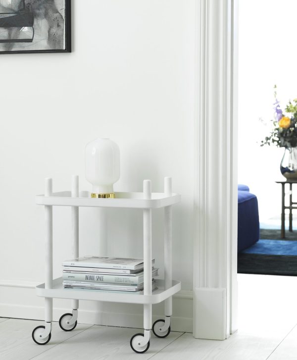 Et lille, hvidt NORMANN COPENHAGEN - Rullebord med to hylder med bøger og en lampe, placeret i hjørnet af et lyst rum nær en døråbning, med et glimt af en blå sofa i det tilstødende rum.