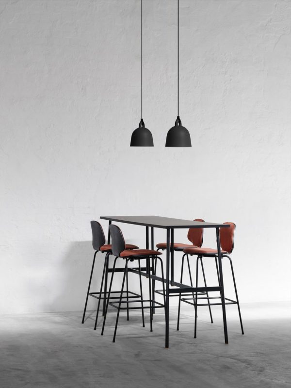 En minimalistisk spiseplads med en NORMANN COPENHAGEN - Bell Lampe Ø22 og fire taburetter med røde sæder under to hængende sorte pendellamper, mod en almindelig hvid væg og et gråt gulv.