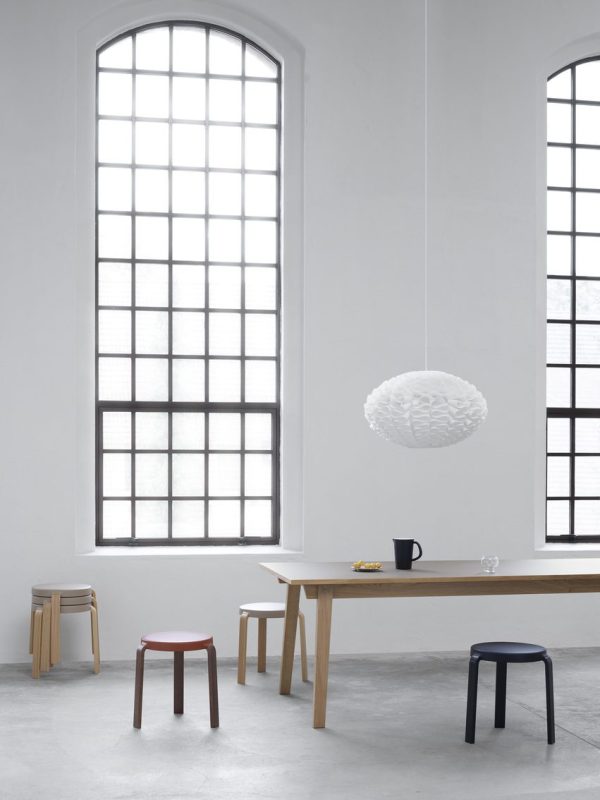 En minimalistisk spiseplads med et træbord, to sorte og en pink skammel. En moderne hvid pendel, der viser TomorrowsDesign, hænger ovenover. Rummet har store buede vinduer og grå vægge.