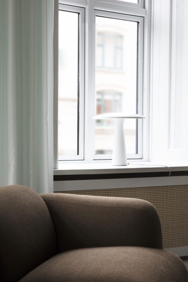 Et moderne værelseshjørne, der viser en del af en Normann Copenhagen - Swell Sofa 3 Pers, en enkel hvid vase i hvid vindueskarm, lyse gardiner og udsigt til en bygning uden for vinduet.