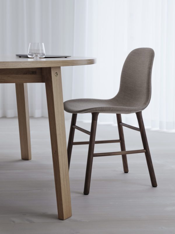 En moderne spiseplads med et træbord og en stofpolstret stol i et minimalistisk værelse med gennemsigtige gardiner og et lyst trægulv. Bordet, der viser TomorrowsDesign, er dækket med et klart glas og en tallerken.