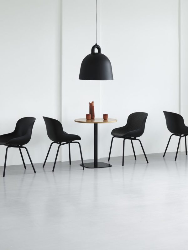 Et minimalistisk interiør med et rundt bord med to stearinlys på, omgivet af fire sorte stole, under en stor sort pendel i et rum med hvide vægge og gråt gulv.