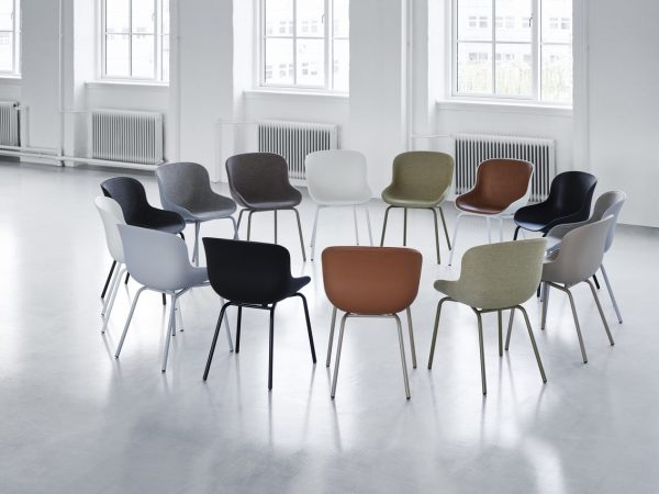 En række moderne stole med forskellige farver og designs arrangeret tilfældigt på et skinnende hvidt gulv i et lyst rum med store vinduer.