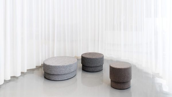 Tre NORMANN COPENHAGEN Silo Puf Medier af varierende størrelser med forskelligt tekstureret gråt polstring, arrangeret på et skinnende gulv mod en baggrund af flydende hvide gardiner.