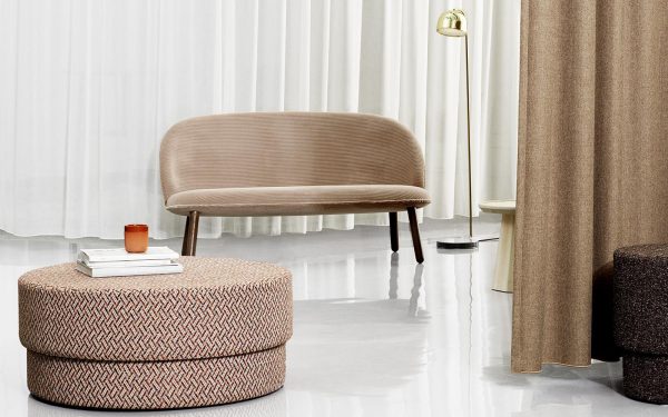 Interiør med en beige afrundet NORMANN COPENHAGEN - Silo Puf Stor, en vævet cirkulær skammel, en lille stak bøger med et rødt glas på toppen og elegante gulvlamper, sat mod en baggrund af lyse gardiner og reflekterende gulve.