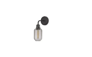 En moderne vægmonteret lampe med NORMANN COPENHAGEN - Amp Væglampe, en matsort cirkulær plade med en enkelt synlig pære indkapslet i en klar, aflang glasskærm, alt sammen mod en almindelig hvid baggrund.