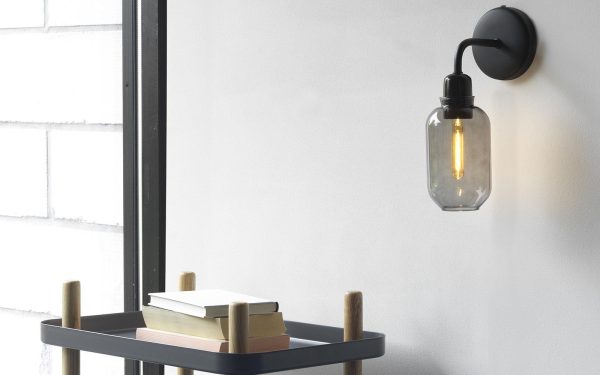 Et minimalistisk interiør med en Normann Copenhagen - Amp Væglampe med en synlig edison-pære ved siden af et vindue. en lille, sortrammet vogn holder pænt stablede bøger under lyset.