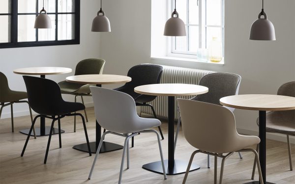 Et moderne caféinteriør med flere runde borde parret med diverse grå og grønne stole med eksklusive designmøbler. Store vinduer tillader naturligt lys at oplyse rummet, og der hænger pendellamper over hvert bord.