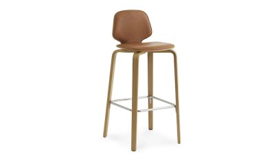 En moderne barstol med karamelbrunt lædersæde og ryglæn, understøttet af slanke gyldne metalben og med fodstøtte. Denne TomorrowsDesign skammel er sat mod en almindelig hvid baggrund.