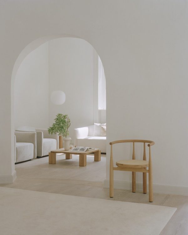 En minimalistisk stue med en buet døråbning, beige vægge, et lyst trægulv og enkle møbler, herunder en træstol, et sofabord og et NYT VÆRK - Atlas Sofabord 82x82 nær et vindue. Blødt naturligt lys fylder rummet.