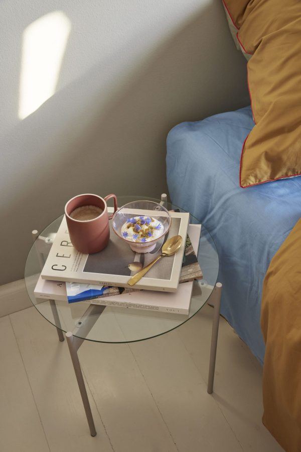 Et hyggehjørne med et sidebord i glas fra TomorrowsDesign, med en skål korn, en rød kop med en drik og magasiner. Sollys kaster en skarp skygge på væggen. En blå sofa med et gult tæppe er delvist synligt.