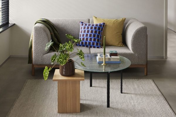 En moderne stue med en grå to-personers sofa med farverige puder, et glas sofabord med bøger og en potteplante og ekstra grønt ved siden af sofaen. Dette rum repræsenterer perfekt TomorrowsDesign-etoset.