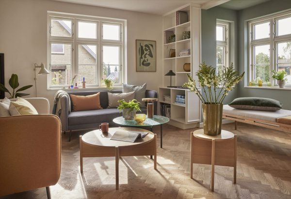 En lys, moderne stue designet af TomorrowsDesign med sildebenstrægulve, med stilfulde møbler, store vinduer, planter og blødt naturligt lys. Indretningen omfatter en sofa, stole og en bogreol.
