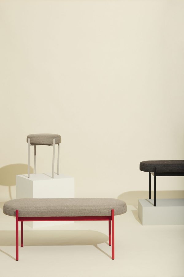 En minimalistisk udstilling af tre moderne møblerbænke med polstrede sæder, hver med særskilte rammedesign og farver, kunstnerisk arrangeret på hvide platforme mod en bleg beige baggrund.