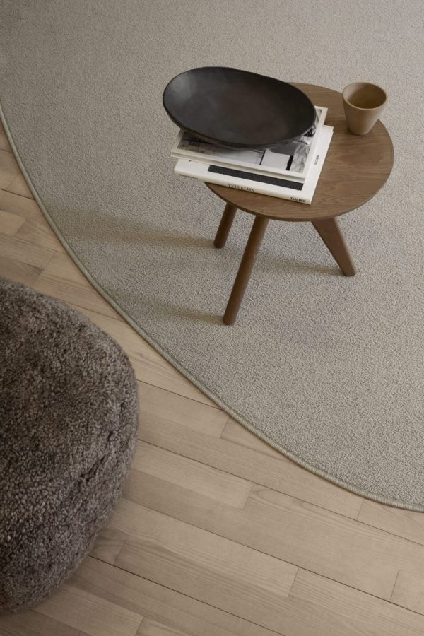 En minimalistisk stue med et cirkulært træsidebord med en sort oval bakke og bøger på toppen, sat på et beige tæppe, der delvist overlapper et mørkere trægulv. Et hjørne af et gråt shaggy tæppe er også synligt, suppleret med slanke designmøbler.