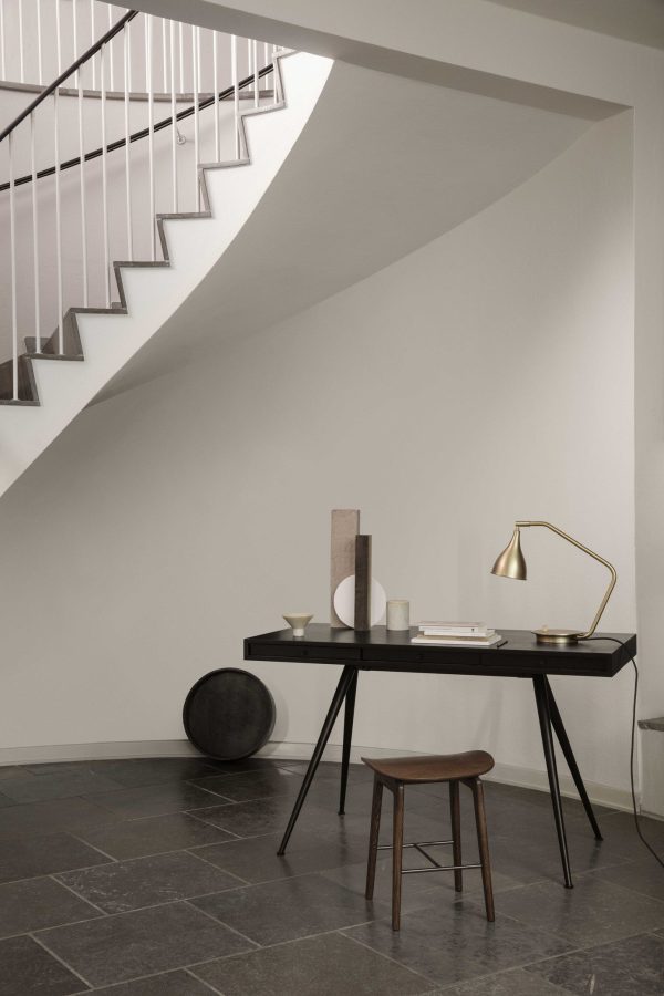 En minimalistisk hjemmekontoropstilling under en hvid trappe, med en NORR11 - Le Six Bordlampe på et slankt, mørkt bord med bøger, dekorationer og et rundt sort dekorationsstykke. En skammel er placeret ved bordet.