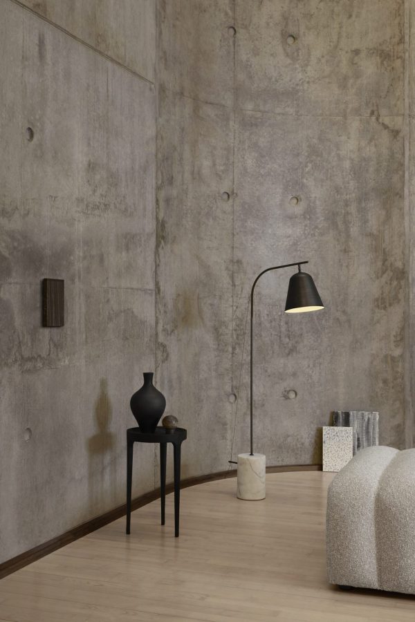 Et minimalistisk værelse med nøgne betonvægge og et hårdttræsgulv, med en NORR11 Line One Gulvlampe, et lille sort sidebord med to vaser og en tekstureret hvid skammel.