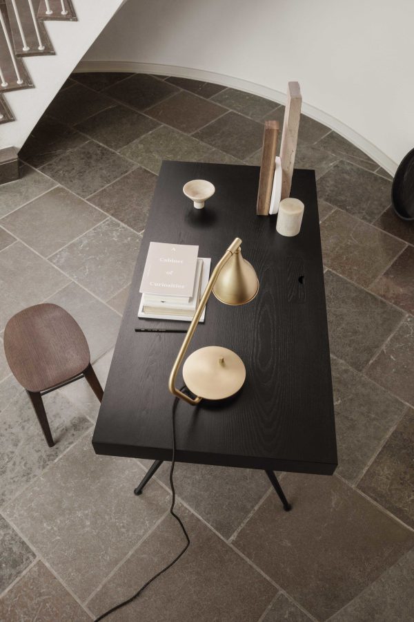 Et moderne værelse med et bord med sort tekstur med en NORR11 - Le Six Bordlampe, bøger og dekorative genstande. en træstol og geometrisk mønstrede trapper er delvist synlige.
