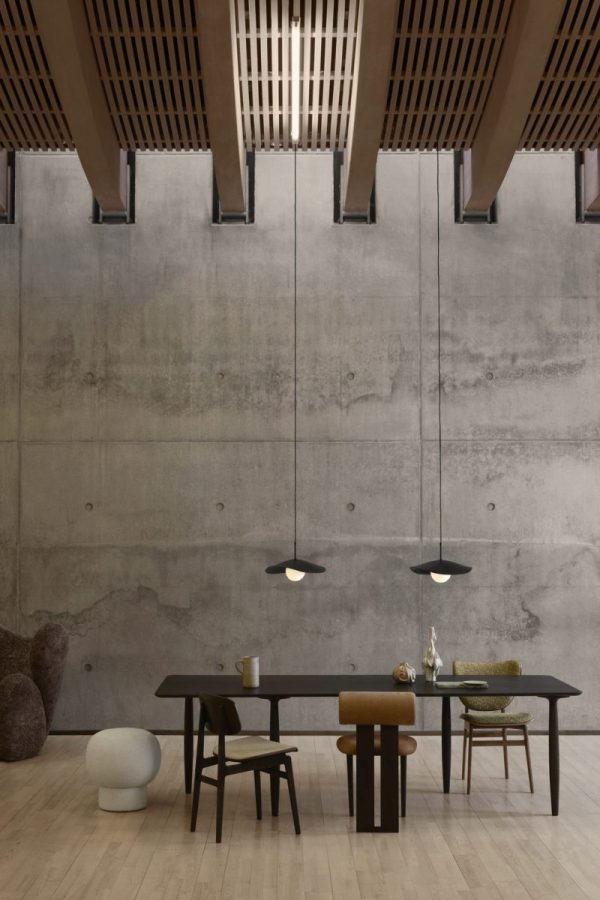 En minimalistisk spisestue med et langt bord med diverse stole, geometriske pendellamper og en rå betonvæg. Et tekstureret træloft tilføjer varme til rummet og inkarnerer essensen af TomorrowsDesign.