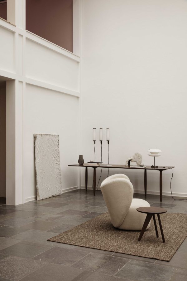 Værelse i minimalistisk stil med et enkelt bord med en skulptur, tre lysestager og en lampe, en polstret stol foran og et indrammet kunstværk lænet op ad væggen. Interiøret lægger vægt på rene linjer og ryddelige rum.